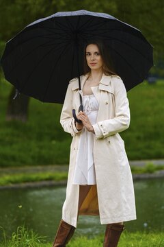 Woman stands by water in park with black umbrella in rain. She wears light dress and light coat. Trees are behind her
