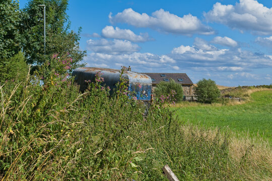 A rural landscape features a rusty blue trailer partially obscured by green plants. Beyond, farm buildings and fields stretch under a cloudy sky - Church Wood and Old Quarry, Bury, UK