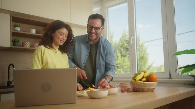 Smiling couple preparing fresh fruit salad with laptop recipe at home kitchen