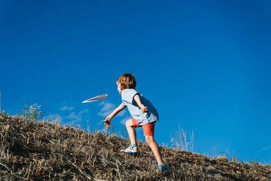 A happy young boy throws a white paper airplane into the air while standing on a grassy hill. Carefree outdoor playtime and joyful childhood concept.