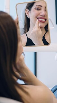 Over the shoulder shot of a happy woman looking at her reflection in a hand mirror, admiring the result of a dental treatment. Vertical
