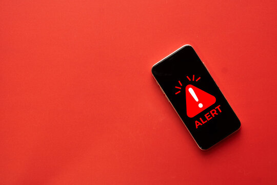 Emergency alert and warning notification concept. Smartphone displaying a red caution triangle and ALERT text on screen against a solid red background with copy space.