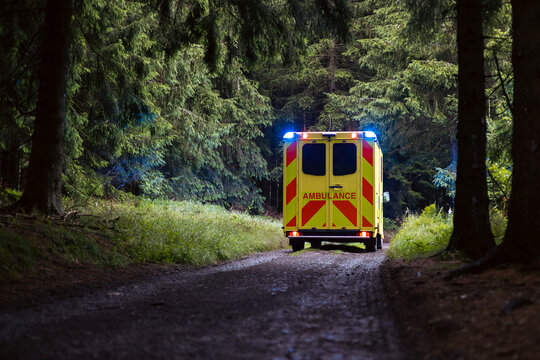 Emergency medical service ambulance vehicle driving on narrow rural road inside dense forest at twilight.  Rescue in remote location, urgent healthcare response and medical transport concept.