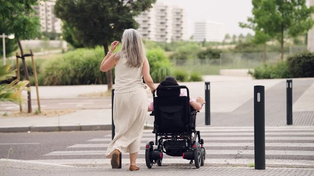 Rear view of a gray haired woman helping her disabled daughter in a motorized wheelchair cross a street on a crosswalk in the city