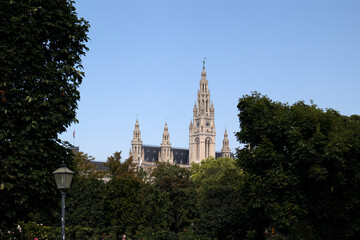 Obraz premium La maestosa torre neogotica del Municipio di Vienna Rathaus che svetta sopra gli alberi del parco Rathauspark con passanti lungo il sentiero in una giornata estiva, Austria.
