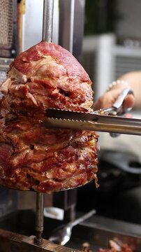 Chef cutting roasted meat from vertical rotisserie grill machine with tongs at local Asian street food market stall. Close up of preparing doner kebab and shawarma, fast food concept.