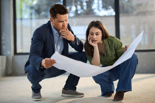 Worried couple cooperating while reading blueprints of their developing house. 