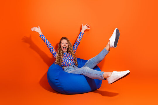 Joyful young woman sitting on a blue beanbag with arms raised against a vibrant orange background