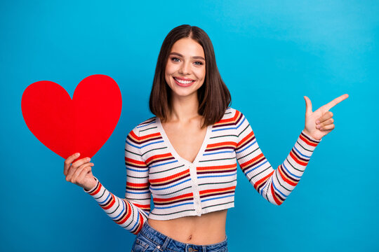 Young woman holds a red heart and smiles wearing a striped cropped cardigan against a blue background for love fashion lifestyle and shopping promotion