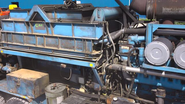 Truck mounted diesel engine assembly in industrial yard with shifting sunlight across blue pipes, twin air filters, metal cab details, rust, paint wear, and maintenance textures