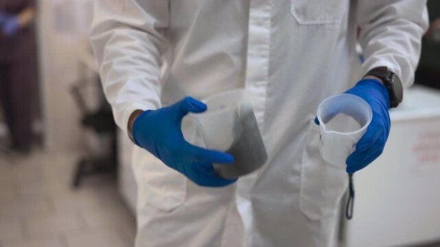 Laboratory technician comparing sample materials by hand with measuring cups in close-up scene, wearing protective gloves and lab coat beside dense gray compound and empty container
