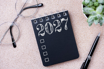 Fototapeta premium New year goals 2027. 2027 resolutions list with notebook, coffee cup, plant on table. Resolutions, plan, goals, action, checklist, idea concept. New Year 2027 resolutions, copy space