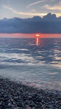 low sun sets beyond a calm sea viewed from a pebble beach, while vivid red reflections shimmer across rolling water beneath large dramatic clouds at late summer dusk