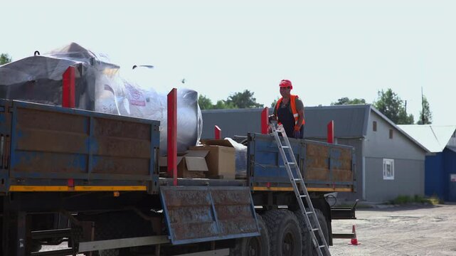 Worker standing on loaded flatbed truck beside ladder in bright industrial yard with covered cargo, cardboard boxes, weathered metal sides, warehouse buildings, dusty ground, pale sky, and distant