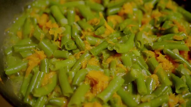 A close-up view of fresh chopped green beans and grated carrots simmering in a frying pan. Concept of healthy vegan diets, authentic home cooking, and real time preparation.