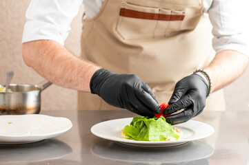 A professional chef wearing black gloves carefully plates a gourmet dish with green crepes and a...