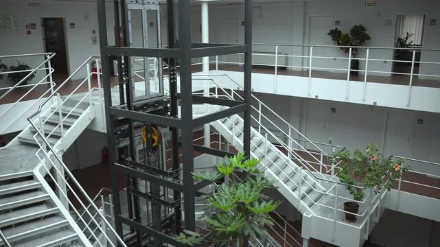 Interior atrium with exposed elevator shaft, visible pulley system, white staircases, mezzanine walkways, stainless handrails and industrial details inside modern institutional building