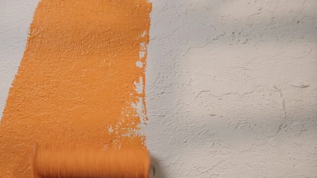 Hand with roller in orange color paint repainting white textured wall in room close-up, dramatically changing interior and atmosphere of house, renovation and improvement at home, mood swings