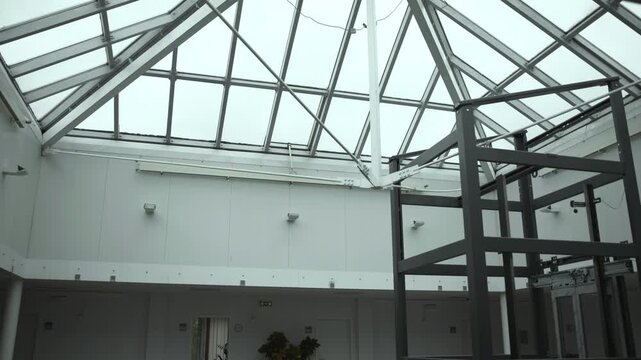 Industrial atrium with pitched glass skylight, triangular steel trusses, diagonal braces, and foreground metal framework viewed from low angle with cool diffuse daylight