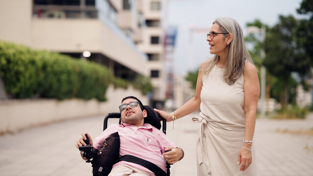 Woman supporting and accompanying a man with spina bifida in an electric wheelchair, showing assistance and inclusion outdoors