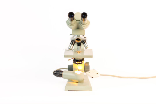 Front view of a classic binocular laboratory microscope with illuminated base and objective lenses. Scientific optical instrument isolated on a white background with ample copy space.