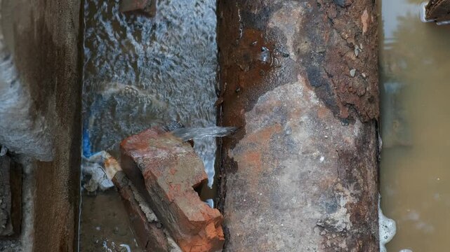 Macro close up view of high pressure water spraying from broken hole in rusted steel utility pipe onto brick wall in trench pit.