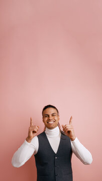 Smiling young Latino man wearing a turtleneck and vest, pointing fingers up, indicating space for text above on a pink background