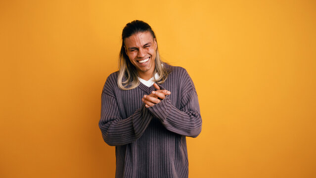 Latino man laughing and smiling with hands clasped, expressing joy and humor against a vibrant yellow studio background
