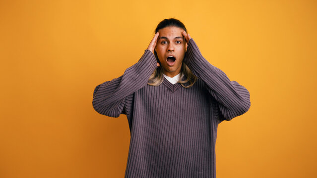 Latino man expressing shock and surprise. He is holding his head with hands, mouth open, standing against an orange background
