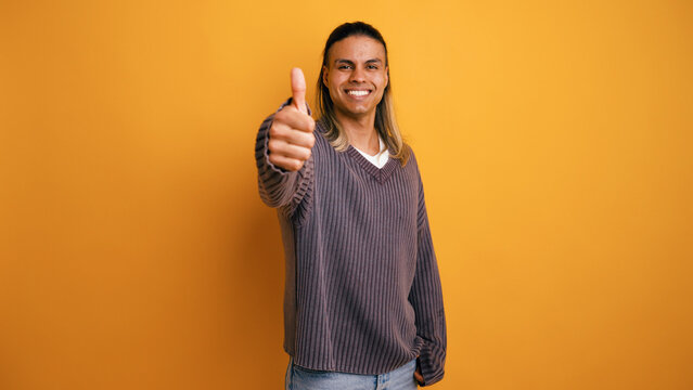 Happy Latino man with long hair smiling and giving a thumbs up, expressing approval and positivity on a yellow background
