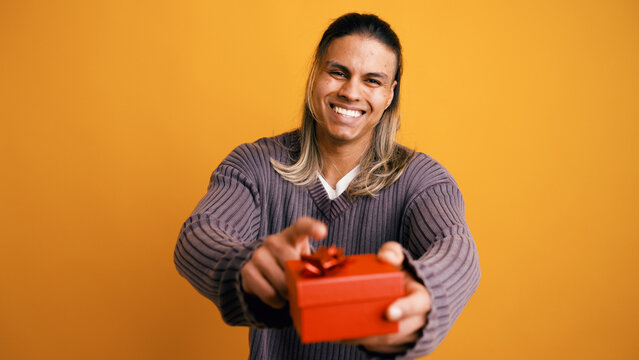 Cheerful latino man offering a wrapped red gift box with a ribbon, celebrating a special occasion in a studio