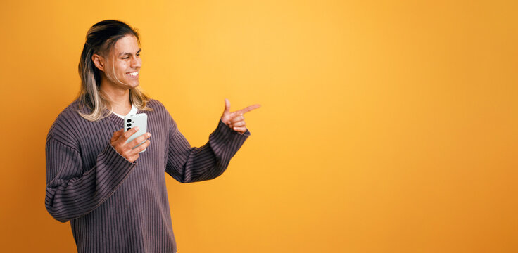 Young latino man smiling, holding smartphone and pointing finger to a blank copy space on orange background