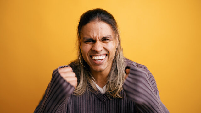 Young Latino man cheering and expressing excitement with clenching fists, celebrating success. Studio shot on orange background