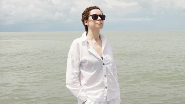 cancer survivor looks toward the camera beside a calm sea, wearing lightweight sun protective clothing for a restorative vacation, resilience and healthy recovery