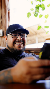 Latino man smiling and relaxing in an outdoor restaurant, wearing glasses and a cap, checking a smartphone