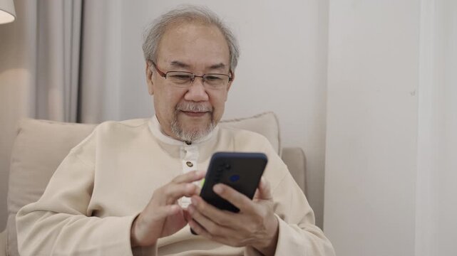 Happy Asian senior man in eyeglasses sitting smiling and using smartphone online social media, chat, shopping on sofa in living room. Old men use technology of cellphone read news and messages at home