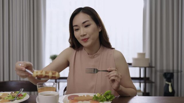 Happy beautiful Asian woman sitting eating breakfast and drinking orange juice for health in the morning in a restaurant at hotel. Attractive young female is having food and drink in kitchen at home. 