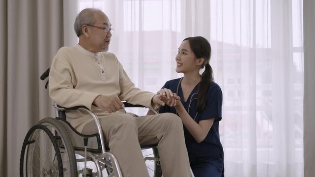 Beautiful young Asian woman nurse caregiver in uniform advises, encourages, and take care of a senior man patient paralyzed with a problem at legs while sitting in a wheelchair in a hospital room. 
