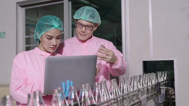 Two Asian quality control inspectors in uniform standing, using a laptop computer to analyze and inspect empty glass beverage bottles on moving conveyor belt in production line at industrial factory. 