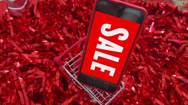 Online sale concept with smartphone in a mini shopping cart rotating on red tinsel. Digital marketing and e commerce for holiday discounts, promotions, and deals