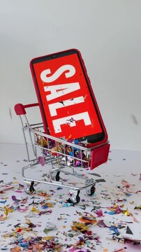 Smartphone with a red screen and white 'sale' text inside a miniature shopping cart while colorful confetti falls from above, isolated on a white background