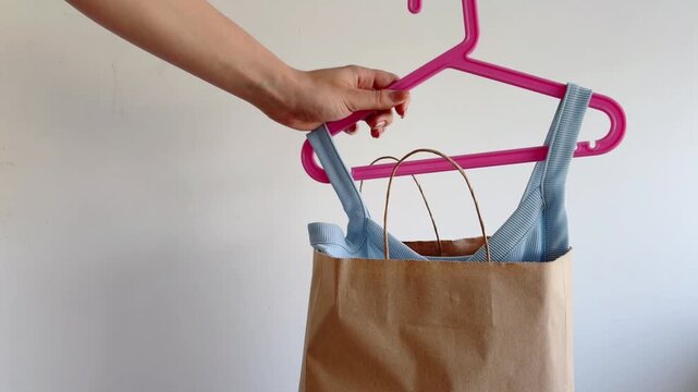 Woman's hand putting new blue clothes on a pink hanger into a brown paper shopping bag after a successful shopping session on a white background, representing consumerism