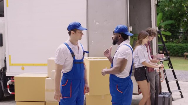 Happy professional movers in uniform standing talking and high fiving after finishing unloading from a truck while a young couple of homeowners are checking and signing for delivery at their new home.