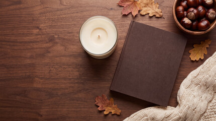 Naklejka premium Autumn comfort concept with book, candle, chestnuts, and warm sweater on dark wood background