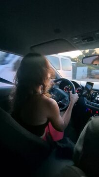 Young woman driving car on residential street, looking out window while steering. Handheld interior shot tracking driver from back passenger seat.
