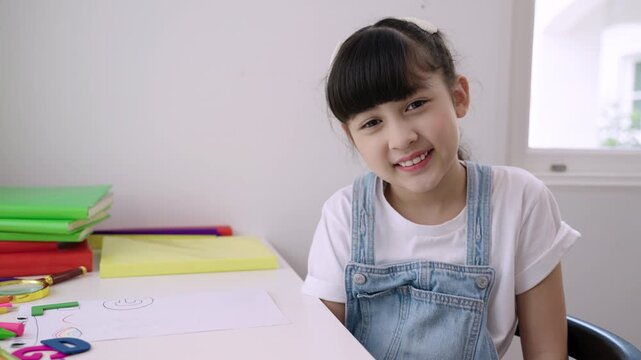 Cute Asian girl student sitting at desk is looking at the webcam while talking to computer camera and video calling with a remote teacher for online learning at home. Homeschooling education concept. 