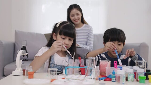 Happy Asian boy and girl students are sitting study learning science and conducting chemistry experiments together with a young teacher sitting and taking care in the back of a nursery classroom lab. 