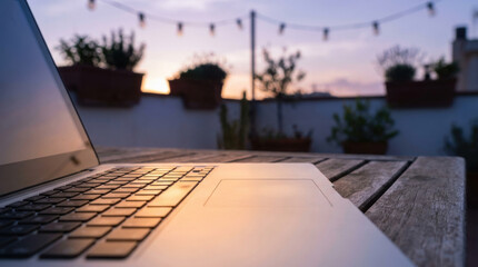 Fototapeta premium Laptop rooftop sunset table keyboard touchpad terrace evening string light wood blurred background outdoor twilight open screen device workspace