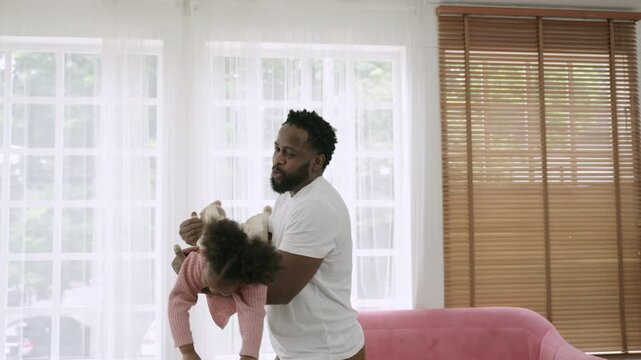 Father's Day concept. Happy African father holding his daughter and playing flying in superhero style, together, having fun and being cheerful in the living room at home. Happy family time and day. 