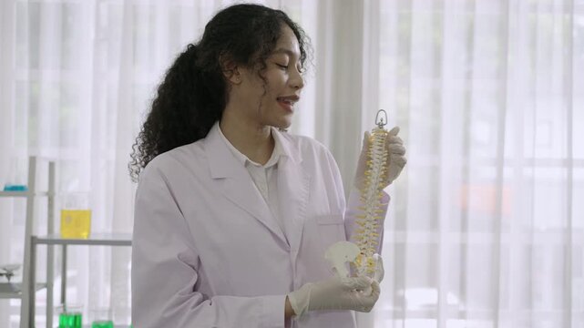 Happy professional woman medical anatomist wearing white lab coat uniform standing holding an anatomical model of the human spine with presentation and explanation carefully detailed in lab at clinic.